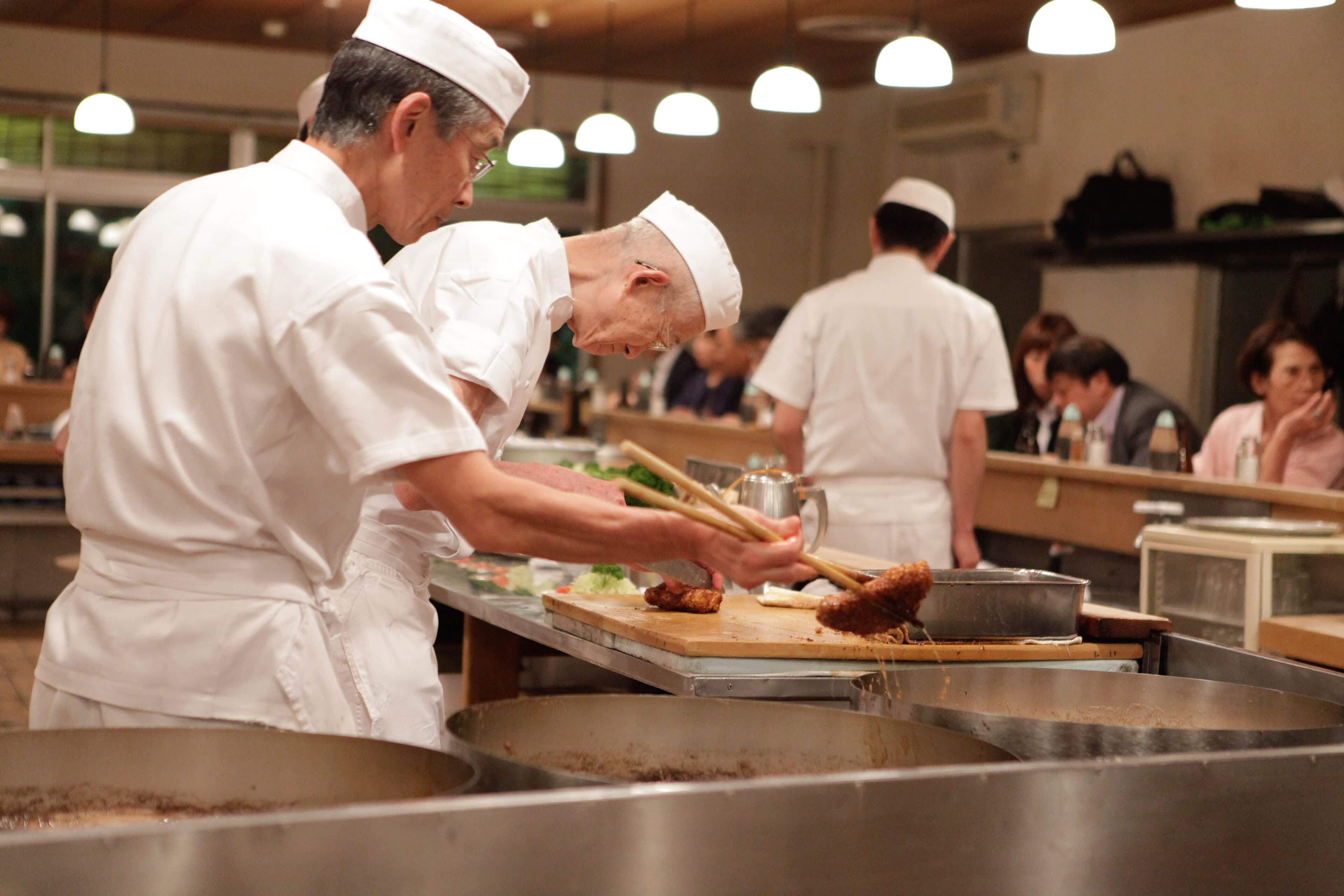 Due cuochi in divisa bianca al lavoro dietro il bancone di un ristorante, con le pentole in primo piano e i clienti seduti sullo sfondo